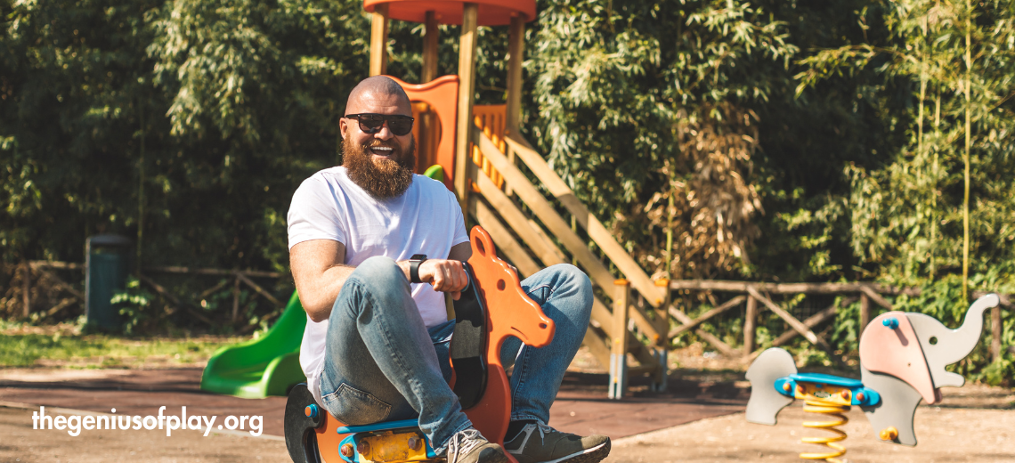 playful man smiling while riding a wooden play horse in a playground 